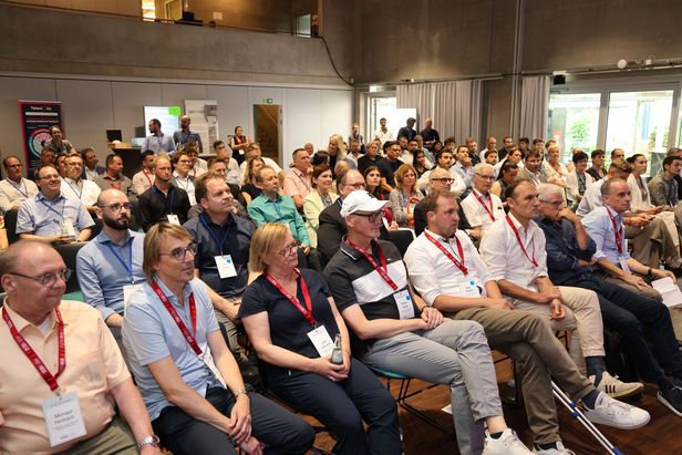Teilnehmende mit Namensschildern sitzen in Reihen in einem Konferenzraum mit Betonwänden und großen Fenstern.