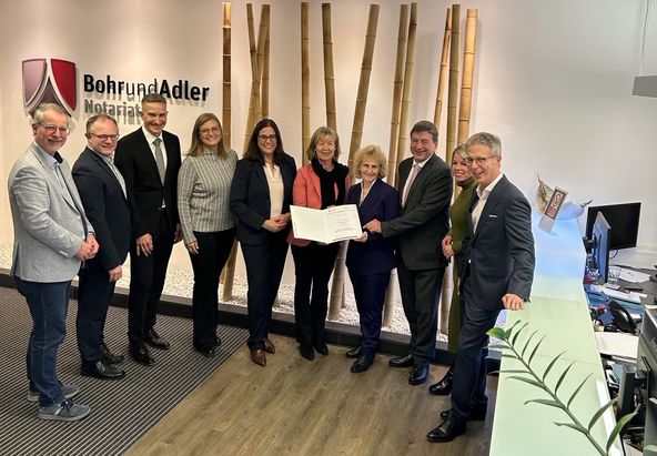 Eine Gruppe von Personen steht in einem modernen B&uuml;robereich vor einer Wand mit Bambusdekor und dem Logo Bohr und Adler Notariat. In der Mitte h&auml;lt die Gruppe ein ge&ouml;ffnetes Dokument. Rechts im Bild befindet sich ein Empfangstresen mit einem Monitor und einer Pflanze.