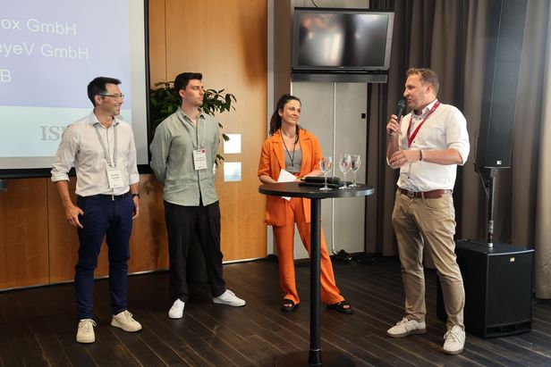 Vier Personen stehen auf einer Bühne an einem Stehtisch mit Mikrofonen und Gläsern und sprechen im Rahmen einer Diskussionsrunde.