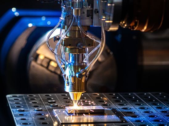 Laserauftragsschweißanlage bearbeitet eine Metallplatte. Aus der Bearbeitungseinheit treten helle Funken aus, während Metallpulver und Laserstrahlen eine Schutzschicht auf der Oberfläche erzeugen. Die Nahaufnahme zeigt den industriellen Beschichtungsprozess in einer Produktions- oder Forschungsumgebung.