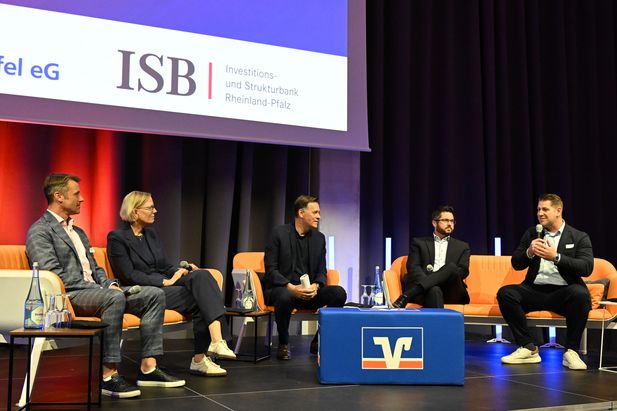 Podiumsdiskussion mit f&uuml;nf Teilnehmenden auf einer B&uuml;hne. Im Hintergrund sind die Logos von ISB und VR Bank RheinAhrEifel auf einer Leinwand zu sehen