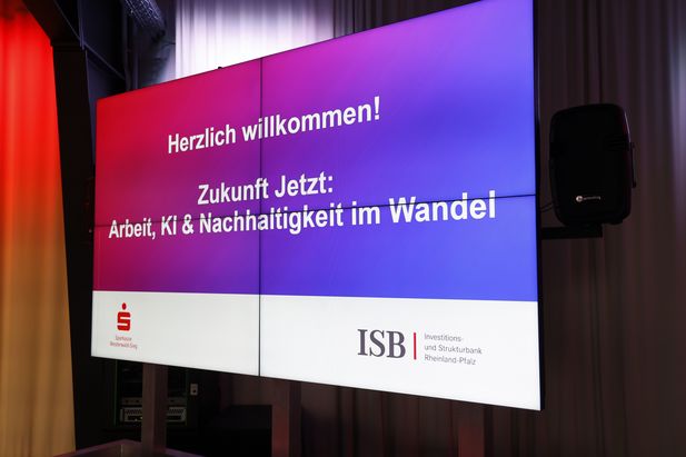 Großer Bildschirm mit der Begrüßungsnachricht „Herzlich willkommen! Zukunft Jetzt: Arbeit, KI & Nachhaltigkeit im Wandel“. Unten sind die Logos der Sparkasse Westerwald-Sieg und der Investitions- und Strukturbank Rheinland-Pfalz (ISB) sichtbar.