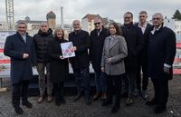 Bescheidübergabe an die Kommunale Wohnungsbaugesellschaft (KWU) in Bad Neuenahr-Ahrweiler: Mehrere Personen stehen in einem Innenraum vor einem Banner. Eine Person überreicht einer anderen ein Dokument, während weitere Personen danebenstehen. Im Hintergrund sind ein Tisch und Informationsmaterial sichtbar.