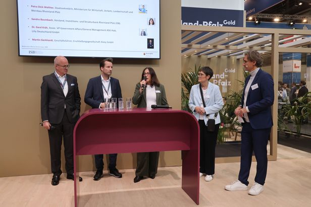 Podiumsgespräch mit fünf Personen vor einem Messestand mit der Aufschrift „Land Pfalz Gold“. Eine Person spricht ins Mikrofon, im Hintergrund zeigt ein Bildschirm Namen und Titel der Redner.