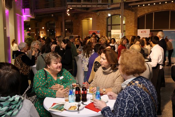 Ein lebhafter Empfangsbereich mit vielen Personen, die an Stehtischen Getränke und Snacks genießen. Auf den Tischen stehen Wasserflaschen, Softdrinks und Teller mit Speisen. Im Hintergrund sind weitere Gruppen im Gespräch sowie Banner verschiedener Organisationen sichtbar. Die Umgebung ist ein heller Raum mit Backsteinwänden und violetten Lichtakzenten.