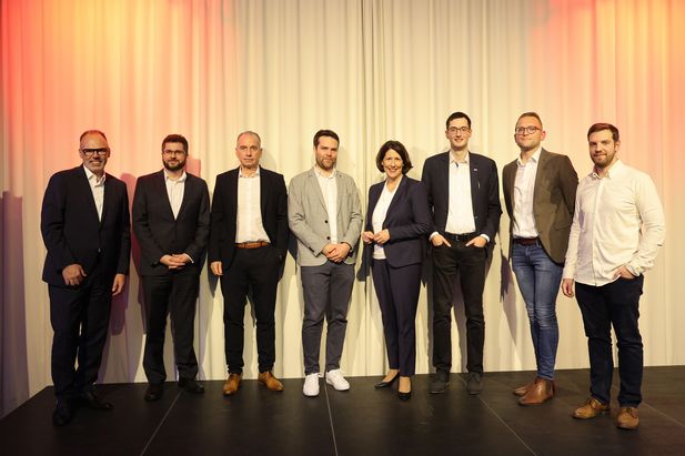 Acht Personen stehen nebeneinander auf einer Bühne vor einem weißen Vorhang. Die Gruppe trägt überwiegend Businesskleidung, bestehend aus Anzügen, Hemden und Blazern. Die Beleuchtung ist warm mit roten Lichtakzenten an den Seiten.