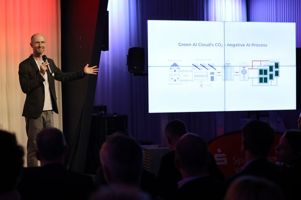 Eine Person steht auf einer Bühne und spricht in ein Mikrofon, während auf einem großen Bildschirm eine Präsentationsfolie mit einem schematischen Diagramm angezeigt wird.