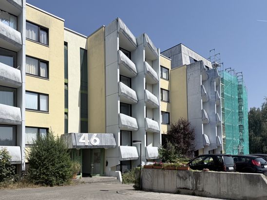 Au&szlig;enansicht eines mehrgeschossigen Studentenwohnheims mit markanten Betonbalkonen und hellgelber Fassade. An einem Geb&auml;udeteil ist ein Bauger&uuml;st mit gr&uuml;nem Netz f&uuml;r laufende Sanierungsarbeiten angebracht.
