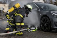 Feuerwehrperson in Schutzkleidung kniet neben einem Elektroauto und bedient einen Schlauch mit angeschlossenem Kühlsystem, aus dem Wasser unter das Fahrzeug und an das Rad gesprüht wird.