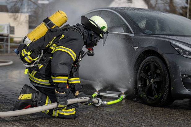Feuerwehrperson in Schutzkleidung kniet neben einem Elektroauto und bedient einen Schlauch mit angeschlossenem K&uuml;hlsystem, aus dem Wasser unter das Fahrzeug und an das Rad gespr&uuml;ht wird.