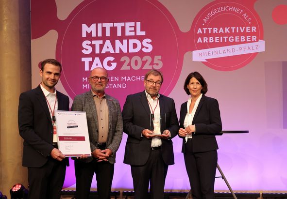 Vier Personen stehen auf einer B&uuml;hne vor einem Hintergrund mit den Aufschriften &bdquo;Mittelstandstag 2025&ldquo; und &bdquo;Attraktiver Arbeitgeber Rheinland‑Pfalz&ldquo;. Eine Person h&auml;lt eine gro&szlig;formatige Urkunde, eine weitere h&auml;lt eine Auszeichnung. Rechts steht ein leerer Stehtisch.