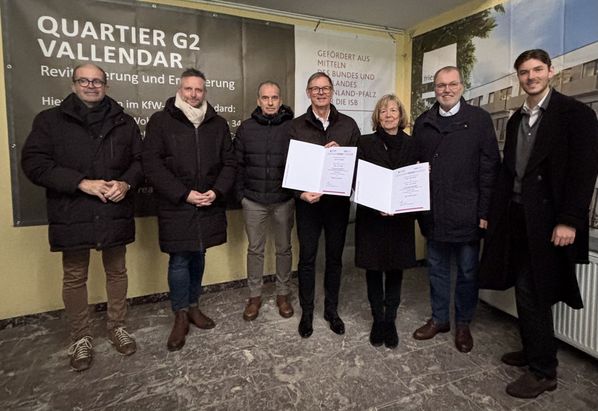 Förderbescheidübergabe für das FRD-Projekt: Mehrere Personen stehen in einem Innenraum vor einem Banner. Eine Person überreicht einer anderen ein Dokument, während weitere Personen danebenstehen. Im Hintergrund sind ein Tisch und Informationsmaterial sichtbar.