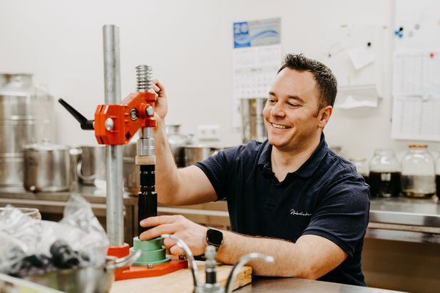 Person an einem Arbeitstisch in einem Produktionsraum bedient ein Messger&auml;t mit roter Halterung. Im Hintergrund befinden sich Edelstahlbeh&auml;lter, Glasgef&auml;&szlig;e und weitere Arbeitsmaterialien.
