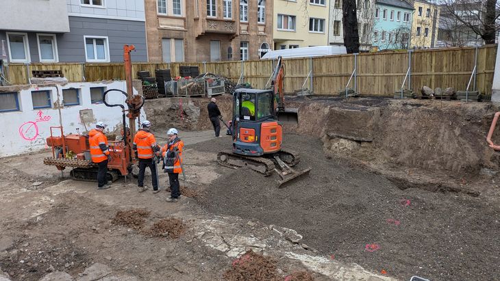 Bauarbeiten in einer ausgehobenen Baugrube: Ein Minibagger und mehrere Fachkr&auml;fte bereiten den Untergrund f&uuml;r den Neubau vor