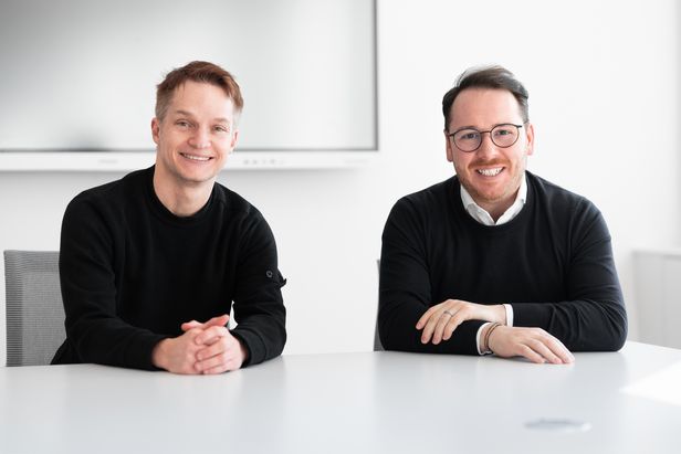 Zwei Personen sitzen nebeneinander an einem wei&szlig;en Konferenztisch in einem hellen Besprechungsraum. Beide tragen dunkle Oberteile, und im Hintergrund ist ein gro&szlig;er Bildschirm an der Wand zu sehen.