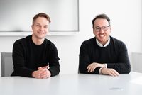 Zwei Personen sitzen nebeneinander an einem weißen Konferenztisch in einem hellen Besprechungsraum. Beide tragen dunkle Oberteile, und im Hintergrund ist ein großer Bildschirm an der Wand zu sehen.