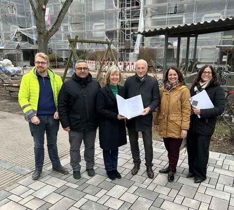 Sechs Personen stehen auf einem gepflasterten Platz vor einem Geb&auml;ude mit Ger&uuml;st und Bauarbeiten. Im Hintergrund sind Bauz&auml;une und ein Baum zu sehen. Zwei Personen in der vorderen Reihe halten ge&ouml;ffnete Mappen mit Dokumenten. Alle tragen winterliche Kleidung, eine Person im hinteren Bereich tr&auml;gt eine auff&auml;llige gelbe Warnjacke.