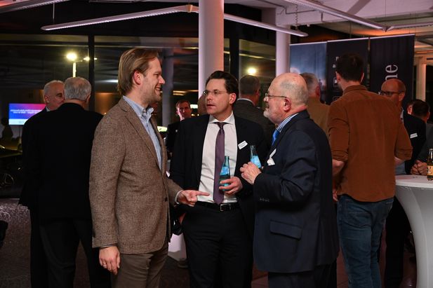 Mehrere Personen stehen in einer Gruppe bei einer Netzwerkveranstaltung und unterhalten sich. Zwei Personen halten Getränkeflaschen, im Hintergrund sind weitere Gäste und Stehtische sichtbar