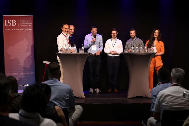 Sechs Personen auf einer Bühne bei einer Podiumsdiskussion hinter zwei Stehtischen mit Mikrofonen und Getränken; links ein Banner mit ‚ISB‘, im Vordergrund das Publikum.