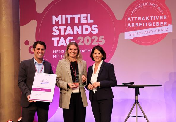 Drei Personen stehen auf einer B&uuml;hne vor einem Hintergrund mit den Aufschriften &bdquo;Mittelstandstag 2025&ldquo; und &bdquo;Attraktiver Arbeitgeber Rheinland-Pfalz&ldquo;. Eine Person h&auml;lt eine Urkunde, eine weitere h&auml;lt eine Auszeichnung. Rechts steht ein leerer Stehtisch.