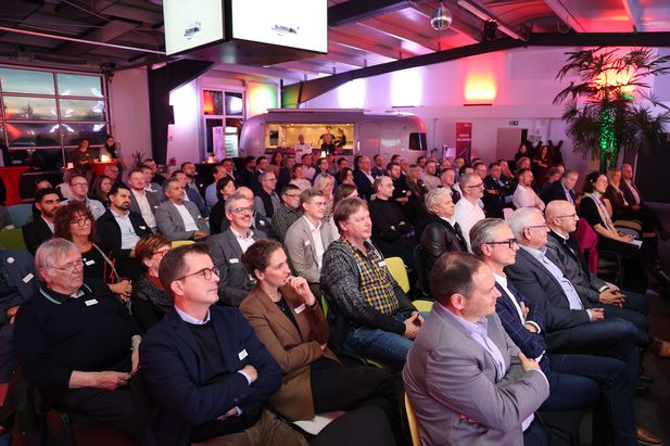 Ein voll besetzter Veranstaltungsraum mit mehreren Reihen von Sitzplätzen. Die Teilnehmenden sitzen und blicken nach vorne in Richtung Bühne. Im Hintergrund sind ein silberner Anhänger, bunte Lichtakzente an den Wänden sowie zwei große Bildschirme an der Decke sichtbar. Rechts steht eine große Pflanze.