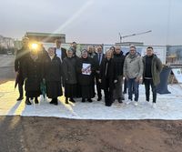 Eine Gruppe von Menschen steht im Freien auf einem hellen Untergrund vor einem Baustellengelände. Eine Person hält ein Dokument in den Händen. Im Hintergrund sind Baucontainer, ein Kran und Informationstafeln zu sehen. Die Gruppe trägt überwiegend wetterfeste Kleidung.