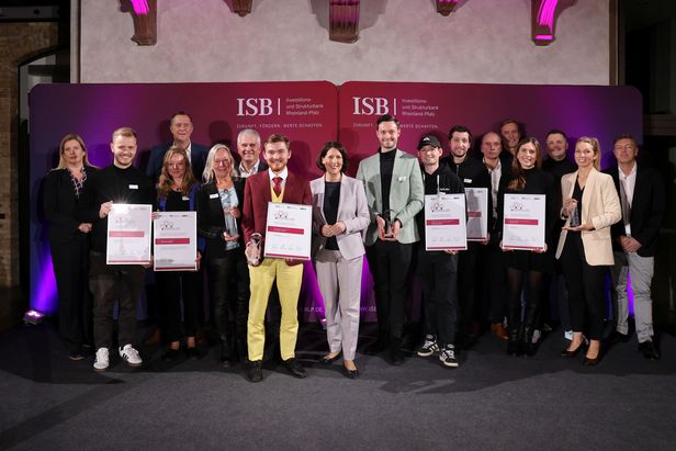 Gruppe von Personen steht vor einer pink-violetten Wand mit dem Logo und Schriftzug der Investitions- und Strukturbank Rheinland-Pfalz (ISB). Mehrere Personen halten Urkunden mit dem Logo des Wettbewerbs ‚Ideenwettbewerb Rheinland-Pfalz‘, andere halten Trophäen.