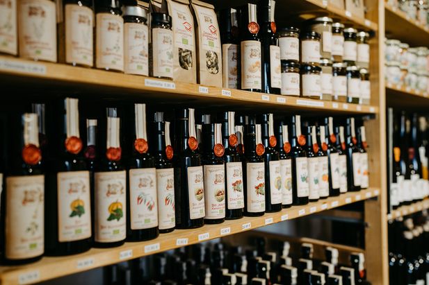 Mehrere Regale mit dunkelgr&uuml;nen und schwarzen Glasflaschen verschiedener Speise&ouml;le sowie weiteren Feinkostprodukten. Die Produkte stehen dicht nebeneinander in Holzregalen und sind mit unterschiedlichen Etiketten und Preisschildern versehen.