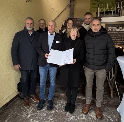 F&ouml;rderbescheid&uuml;bergabe an die Stiftung Bethesda: Mehrere Personen stehen in einem Innenraum vor einem Banner. Eine Person &uuml;berreicht einer anderen ein Dokument, w&auml;hrend weitere Personen danebenstehen. Im Hintergrund sind ein Tisch und Informationsmaterial sichtbar.