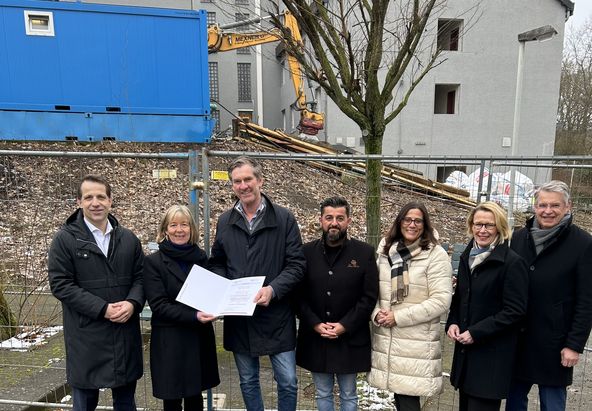 Eine Gruppe von sieben Personen steht im Freien vor einem abgesperrten Baustellengel&auml;nde. Zwei Personen in der Mitte halten gemeinsam ein gr&ouml;&szlig;eres Dokument. Im Hintergrund sind ein teilweise saniertes Geb&auml;ude, ein Baucontainer und ein Bagger zu sehen. Die Gruppe tr&auml;gt wetterfeste Kleidung.
