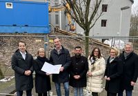 Eine Gruppe von sieben Personen steht im Freien vor einem abgesperrten Baustellengelände. Zwei Personen in der Mitte halten gemeinsam ein größeres Dokument. Im Hintergrund sind ein teilweise saniertes Gebäude, ein Baucontainer und ein Bagger zu sehen. Die Gruppe trägt wetterfeste Kleidung.