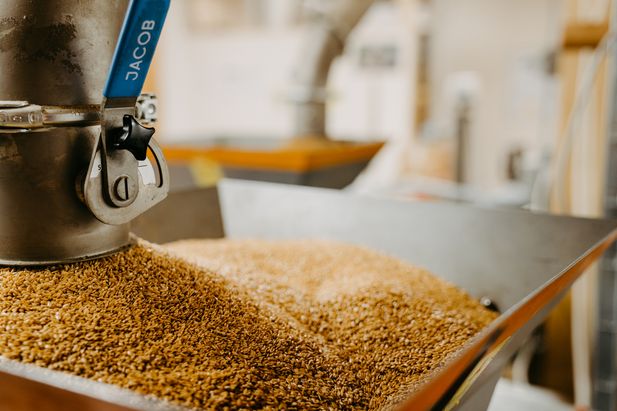 Nahaufnahme eines Edelstahlbeh&auml;lters mit goldbraunen Saaten in einer Produktionsanlage. Links ist ein blauer Hebel an der Anlage sichtbar, im Hintergrund sind weitere technische Einrichtungen unscharf zu erkennen.
