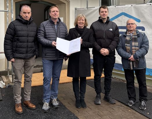 F&ouml;rderbescheid&uuml;bergabe f&uuml;r das Wohnprojekt in Bad H&ouml;nningen: Mehrere Personen stehen in einem Innenraum vor einem gro&szlig;en Banner. Eine Person &uuml;berreicht einem anderen eine Mappe, w&auml;hrend weitere Personen danebenstehen. Im Hintergrund sind ein Tisch und Informationsmaterial sichtbar.