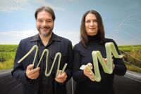 Zwei Personen stehen vor einem Landschaftshintergrund und halten jeweils mehrere geschwungene, helle Kunststoffelemente in den Händen.