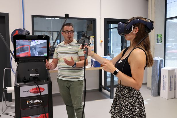 Mädchen rechts im Bild hat eine VR-Brille auf und hält eine digitale Spritzpistole zum Lackieren in der rechten Hand. Auf einem Monitor sieht man das digitale Lackieren.