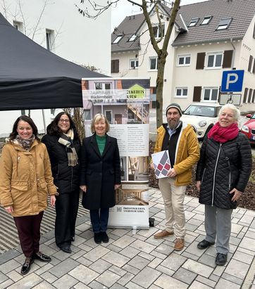 F&uuml;nf Personen stehen auf einem gepflasterten Platz vor einem hellen Geb&auml;ude mit braunen Fensterl&auml;den. Im Hintergrund ist ein schwarzes Zelt und ein Informationsplakat mit Text und Bildern zu sehen. Eine Person in der vorderen Reihe h&auml;lt eine Mappe mit Farbmusterkarten. Alle tragen winterliche Kleidung.
