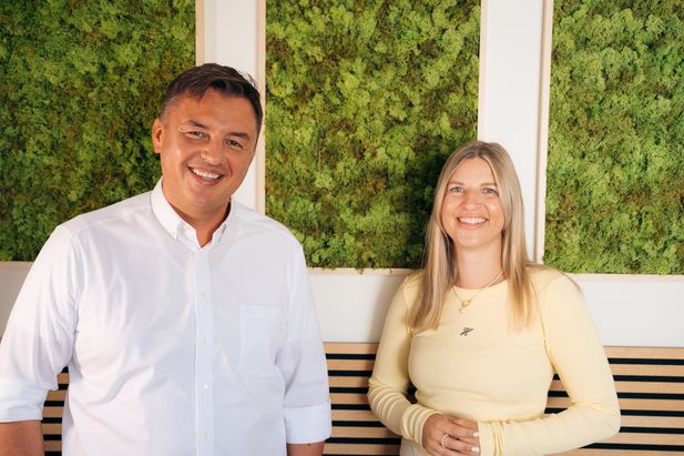 Zwei Personen stehen nebeneinander in einem Innenraum vor einer begrünten Wand; sie tragen helle, gepflegte Kleidung und wirken in einem professionellen Arbeitsumfeld.