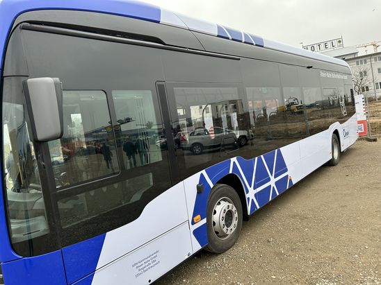 Seitliche Ansicht eines blau‑wei&szlig;en Linienbusses des Nahverkehrsverbunds, der auf einem unbefestigten Baugel&auml;nde steht