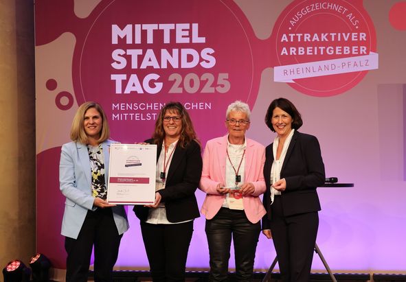 Vier Personen stehen auf einer B&uuml;hne vor einem Hintergrund mit den Aufschriften &bdquo;Mittelstandstag 2025&ldquo; und &bdquo;Attraktiver Arbeitgeber Rheinland‑Pfalz&ldquo;. Eine Person h&auml;lt eine gro&szlig;formatige Urkunde, eine weitere eine Auszeichnung. 