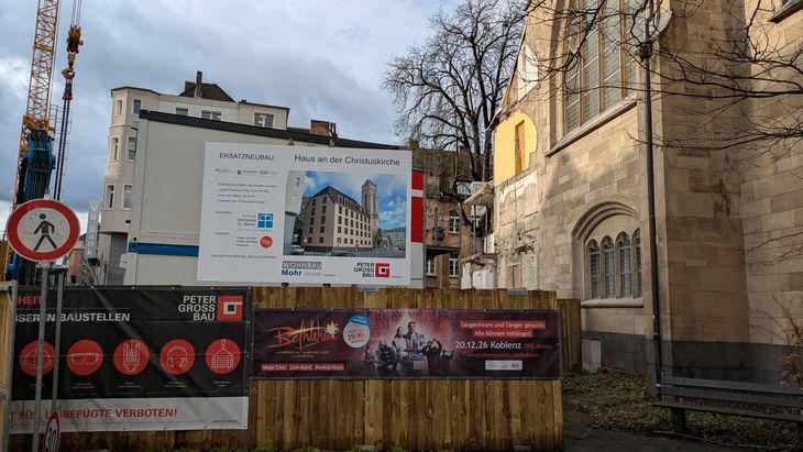 Baustelle neben einer Kirche mit Bauschild des Projekts &sbquo;Haus an der Christuskirche&lsquo;. Das Schild zeigt eine Visualisierung des geplanten Neubaus