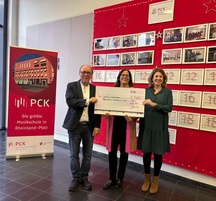 Drei Personen stehen in einem Innenraum vor einer roten Pinnwand mit Fotos und halten gemeinsam einen großen Spendenscheck. Der Scheck zeigt einen Betrag von 3.206 Euro für das Peter-Cornelius-Konservatorium. Links steht ein Roll-up mit der Aufschrift ‚PCK – Die größte Musikschule in Rheinland-Pfalz‘ und einem Foto des Gebäudes.