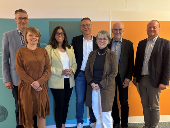 Sieben Personen stehen in zwei Reihen vor einer Wand mit orange  und t&uuml;rkisfarbenen Fl&auml;chen. Die Gruppe tr&auml;gt &uuml;berwiegend business  oder semiformaler Kleidung und blickt l&auml;chelnd in die Kamera.