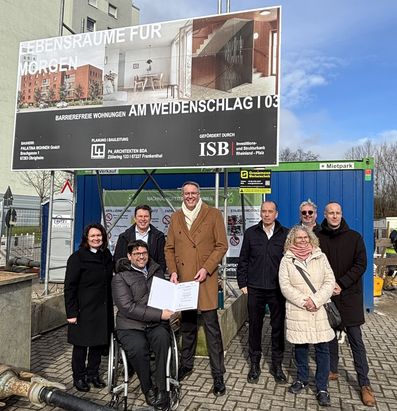 Eine Gruppe von Menschen steht vor einer Baustelle und einem gro&szlig;en Bauschild, das barrierefreie Wohnungen am Weidenschlag ank&uuml;ndigt. Eine Person im Rollstuhl h&auml;lt ein aufgeschlagenes Dokument, w&auml;hrend die anderen gruppiert darum stehen. Hinter ihnen befinden sich ein Container, weitere Hinweistafeln und ein teilweise bew&ouml;lkter Himmel.