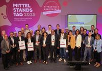 ine große Gruppe von Personen steht auf einer Bühne vor einem farbig gestalteten Hintergrund mit den Aufschriften „Mittelstandstag 2025“, „Attraktiver Arbeitgeber“ und „Rheinland-Pfalz“. Mehrere Personen halten Auszeichnungen oder Urkunden in den Händen. Rechts im Hintergrund ist ein Bildschirm mit dem Titel „Preisverleihung Attraktive Arbeitgeber in Rheinland-Pfalz“ zu sehen.
