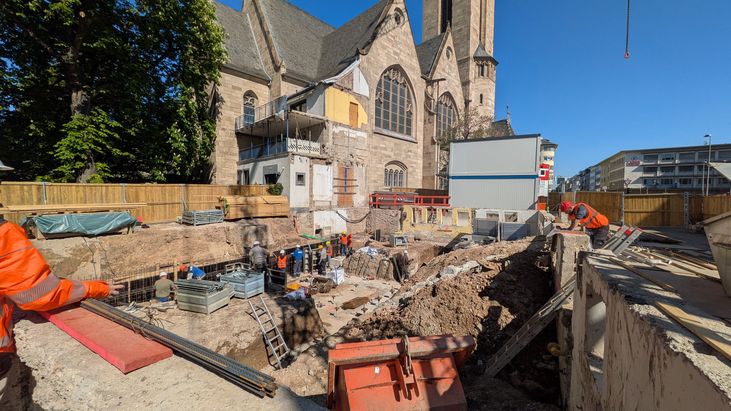 Baugrube mit mehreren Bauarbeitenden und Bewehrungsarbeiten vor einer gro&szlig;en Kirche. Auf dem Gel&auml;nde entstehen Fundamente f&uuml;r einen Neubau