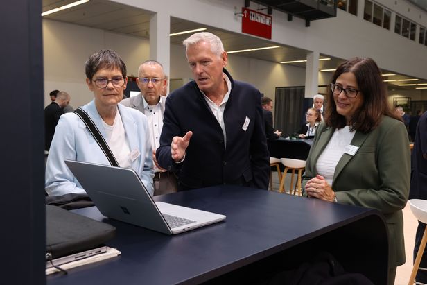 Drei Erwachsene in Businesskleidung stehen um einen Tisch und betrachten gemeinsam einen Laptop-Bildschirm in einem modernen Veranstaltungsraum. Eine Person zeigt auf den Bildschirm und erklärt etwas, während andere aufmerksam zuhören. Im Hintergrund sind weitere Teilnehmer sichtbar.