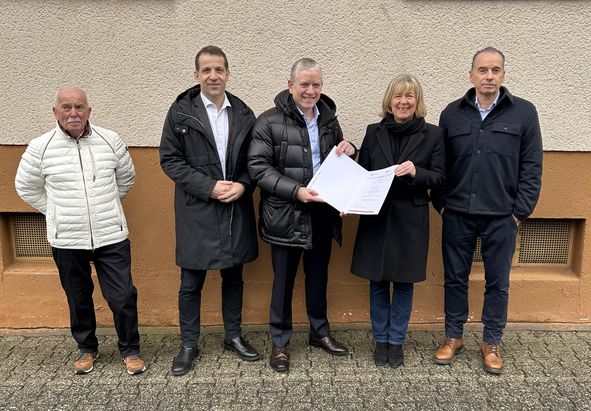 Fünf Personen stehen in einer Reihe vor einem Gebäude mit beigefarbener Wand und weißen Fensterläden. Eine Person in der vorderen Reihe hält ein Dokument, das im Mittelpunkt des Gruppenfotos steht. Alle tragen formelle oder halbformelle Kleidung