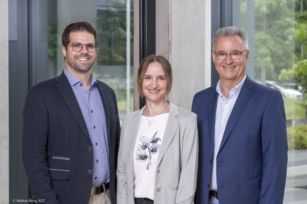 Gruppenportr&auml;t von drei Personen in Gesch&auml;ftskleidung vor gro&szlig;en Fenstern und Betonst&uuml;tzen in einem modernen B&uuml;rogeb&auml;ude.