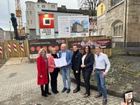 Eine Gruppe von sechs Personen steht vor einer städtischen Baustelle und betrachtet ein aufgeschlagenes Dokument. Im Hintergrund sind Bauzäune, Infotafeln des Unternehmens Peter Gross Bau, ein Kran sowie umliegende Gebäude zu sehen.