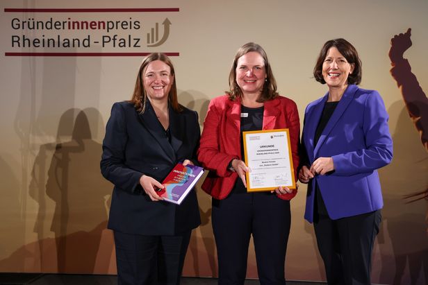 Drei Personen stehen vor einem Banner mit der Aufschrift „Gründerinnenpreis Rheinland-Pfalz“. Eine Person hält ein Buch mit dem Titel „INNOVATIONEN – Zukunft Jetzt“, eine andere eine gerahmte Urkunde. Die dritte Person steht daneben, alle befinden sich auf einer Bühne.